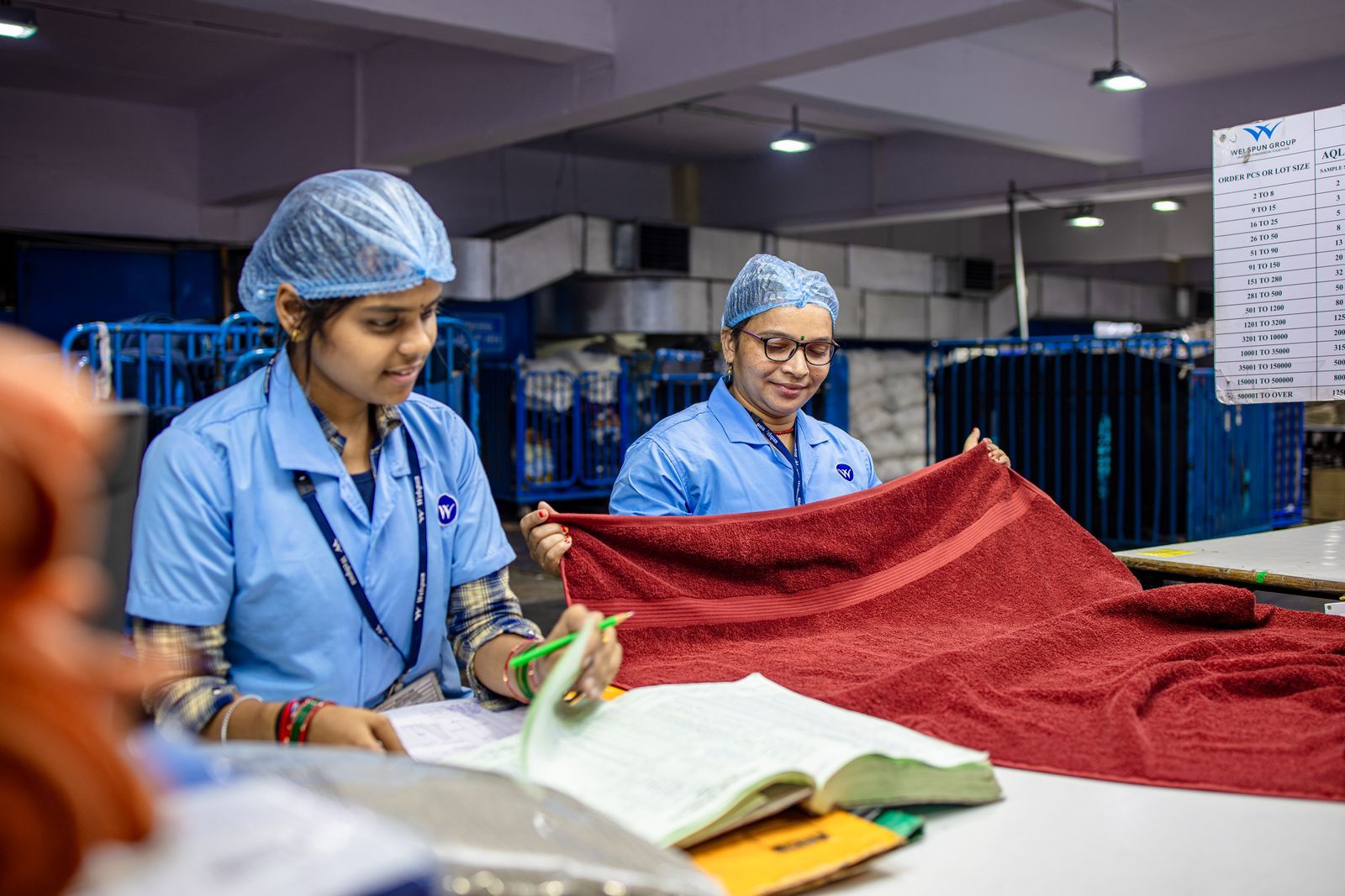 Two quality inspectors examining finished textile goods and recording findings against an AQL checklist at a factory inspection station.